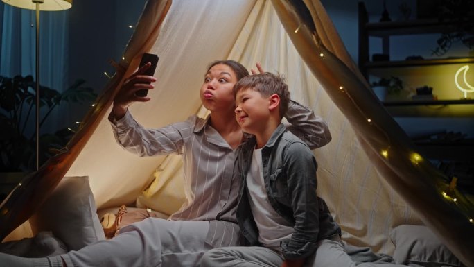 小男孩和他亲爱的妈妈做鬼脸，用智能手机自拍，坐在帐篷里