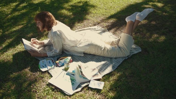 一个受启发的成年女子躺在公园的毯子上，在速写本上画画