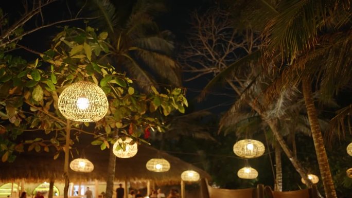 在夜晚的热带背景下，柳条草灯挂在树上作为装饰。老式的木制灯泡在风中摇曳，照亮了户外咖啡馆。波西米亚婚