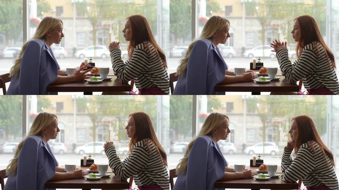 年轻的红发女人和金发的女性朋友聊天的侧视图，在舒适的餐厅一起喝茶，坐在桌子旁