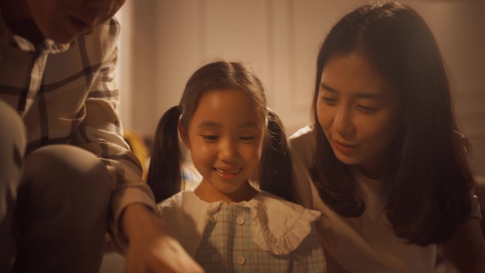 可爱的韩国女孩的特写，在温暖的灯光下，在舒适的公寓里向爱她的父母展示她的画。骄傲的父母支持艺术女儿，
