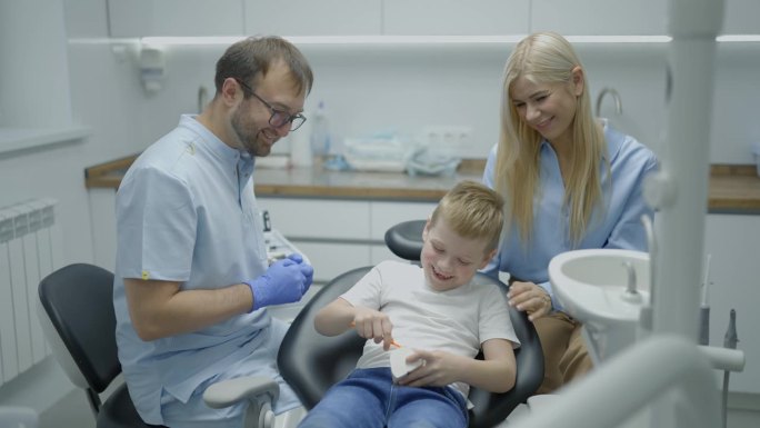 男牙医向男孩和他的妈妈展示下颌模型，教他们如何以有趣的方式正确刷牙。孩子坐在牙科椅上拍着手。慢动作