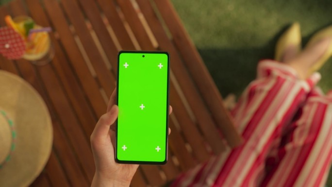 匿名女子在花园里使用智能手机的俯视图，模拟绿屏Chromakey显示屏。优雅女性滚动，网上购物，准备