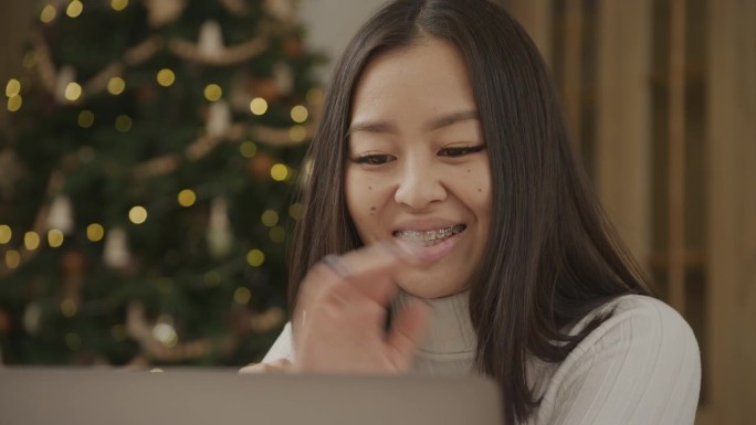 一个亚洲女人用笔记本电脑进行视频通话，室内背景是一棵圣诞树