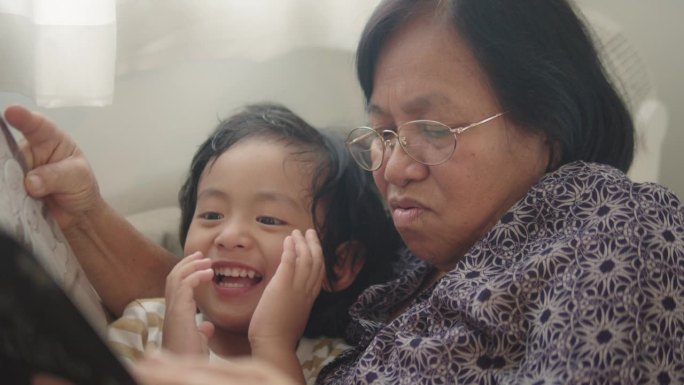亚洲老年妇女穿着舒适的祖母和孙子与她躺在家里的客厅里读故事。