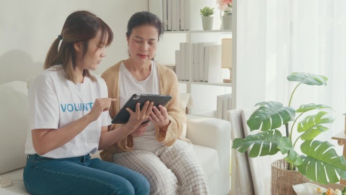 年轻的亚洲女性志愿者教年长的妇女在家里客厅的沙发上使用数码平板电脑。面向老龄化社会的老年素养数字技术