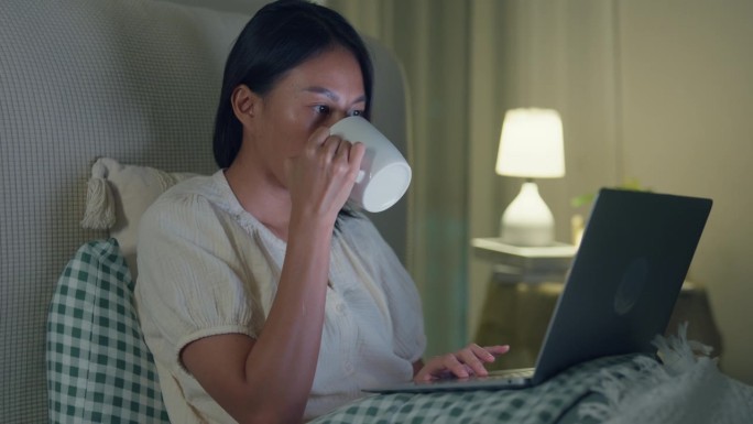 年轻的亚洲女孩晚上坐在家里的床上使用笔记本电脑和喝牛奶。失眠、晕机、无手机恐惧症、睡眠障碍。