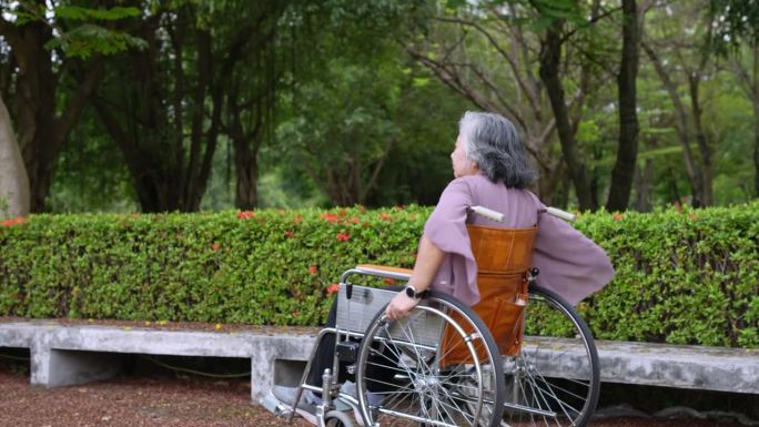 独自快乐的亚洲老年妇女坐在轮椅上，推着轮椅在公园里向前走，在照顾者的照顾下快乐退休的概念，储蓄和老年