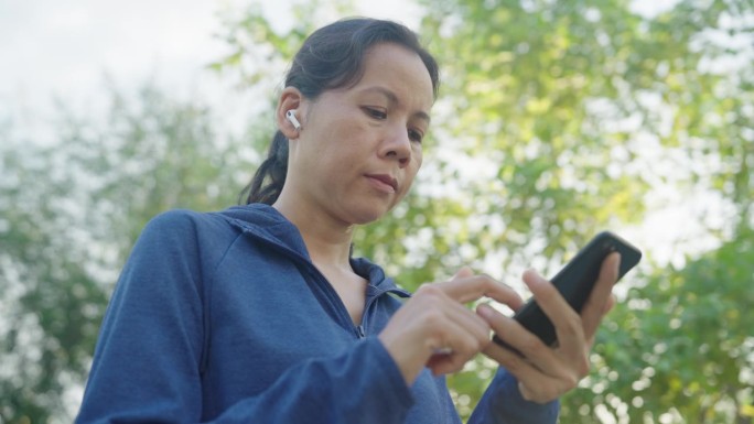 亚洲女运动员穿着运动蓝，站在公园户外用智能手机听音乐，戴着耳机