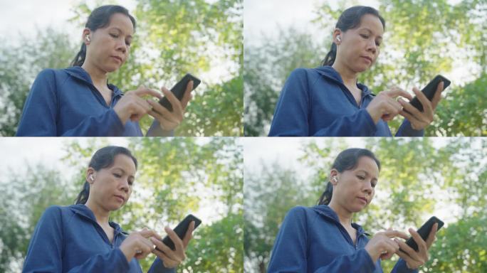 亚洲女运动员穿着运动蓝，站在公园户外用智能手机听音乐，戴着耳机