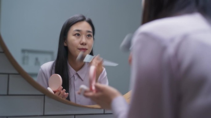 时髦的韩国女性用软毛刷在家里的浴室里涂色调粉底霜。美丽的女人照镜子，在脸上涂上天然的有机化妆品