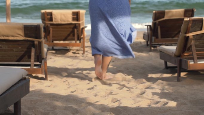 一位金发碧眼、身穿蓝色长裙的年轻漂亮女子沿着海滩散步。穿蓝裙子的女孩。长长的红裙子在风中飘动