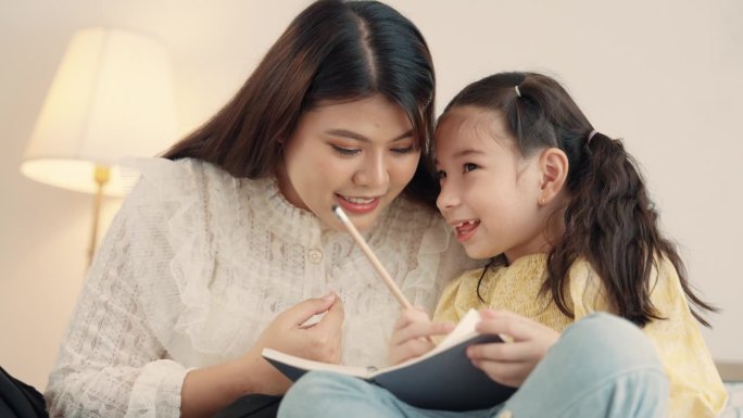 亚洲母亲在家教女儿做作业，在床上玩耍。年轻的母亲在家照顾女儿并教她画画。