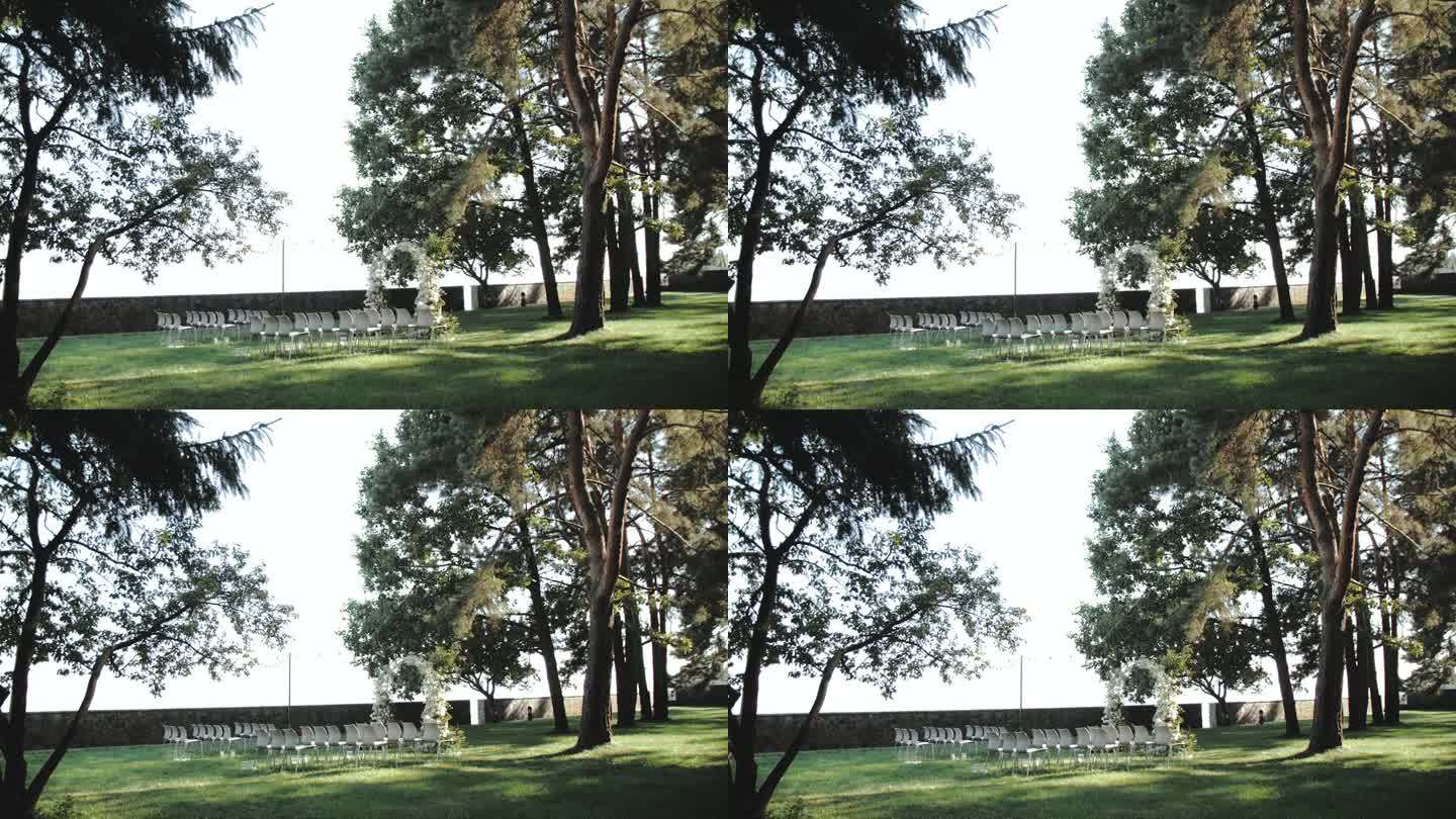 美丽的婚礼拱门装饰着公园里的白色花朵和绿色植物，背景是松树，白色的椅子，阳光洒在草坪上，经典的婚礼，