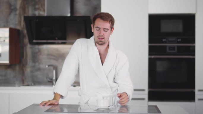 早上穿着浴袍的男人在厨房里打呵欠伸懒腰。实时