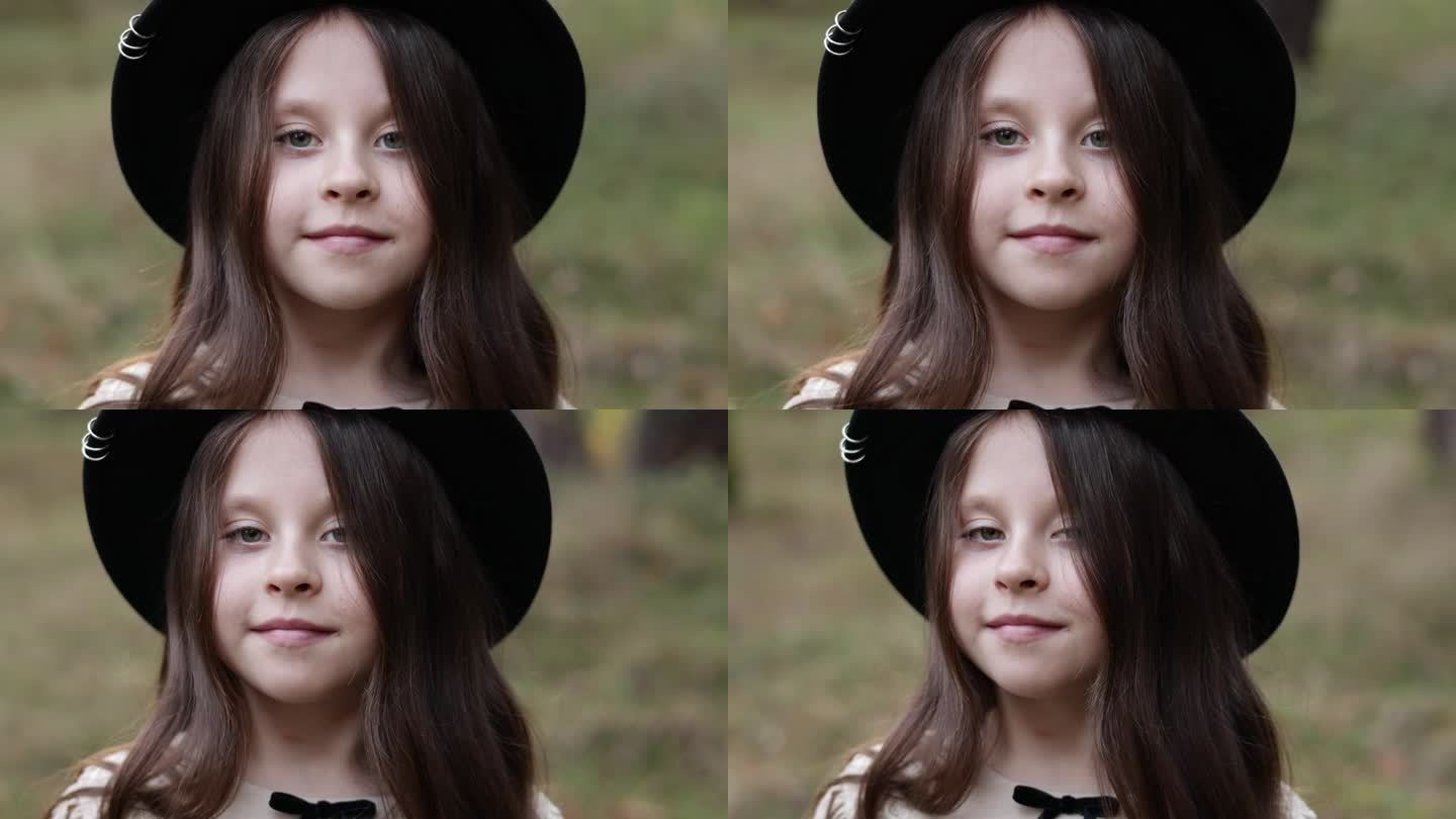 白人小女孩站在户外看着相机的肖像。在绿色公园的背景下，戴着黑帽子的时尚可爱的微笑孩子享受童年的见证概