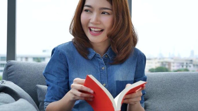 放松的亚洲女人坐在客厅的沙发上拿着书看书。年轻女子放松读书，打开书读书，休闲心灵。幸福美女人笑脸快乐