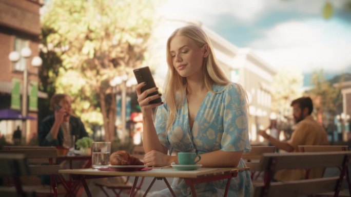 迷人的女人独自坐在街边的咖啡馆里，喝着咖啡，吃着羊角面包。年轻女性使用智能手机，在线与朋友聊天，查看