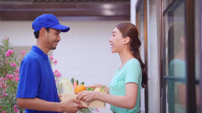 亚洲年轻的快递员在家里给女顾客送包裹。穿着蓝色制服的迷人邮递员正在工作，把一盒水果和蔬菜杂货送到家门