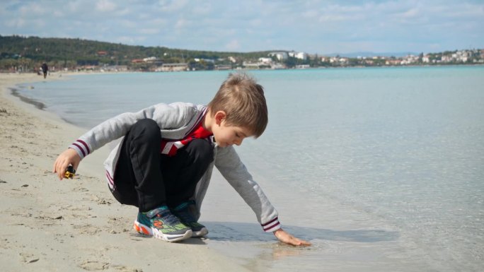 小男孩坐在沙滩上，用手抚摸着冰冷的海水。旅游概念，旅游，暑假。