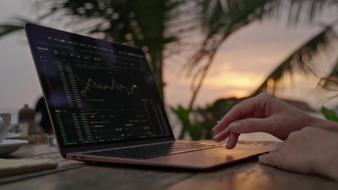 女性加密货币交易员在笔记本电脑上查看在线烛台图，在热带海滨户外咖啡馆远程工作。女经纪人分析汇率和押注