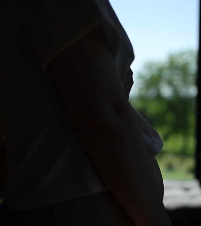 背光下孕妇的肚子。女孩把手放在肚子上，抚摸着肚子。一个孕妇在房子窗户前的剪影