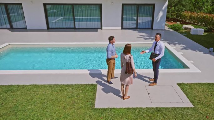 CS男房地产经纪人与一对已婚夫妇握手，他们站在现代住宅的游泳池旁
