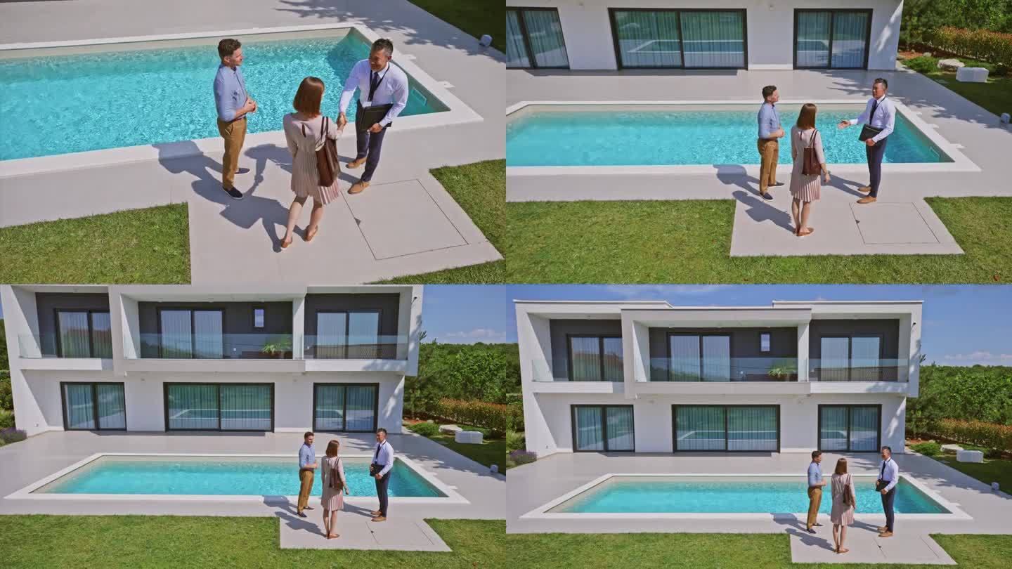 CS男房地产经纪人与一对已婚夫妇握手，他们站在现代住宅的游泳池旁