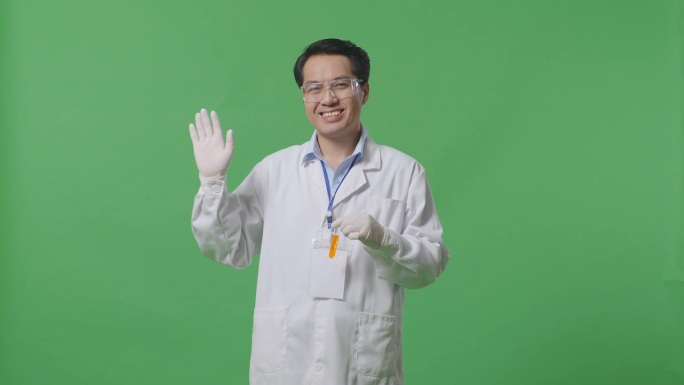 亚洲男性科学家，试管里有橙色液体，站在实验室的绿色背景屏幕上微笑着挥手