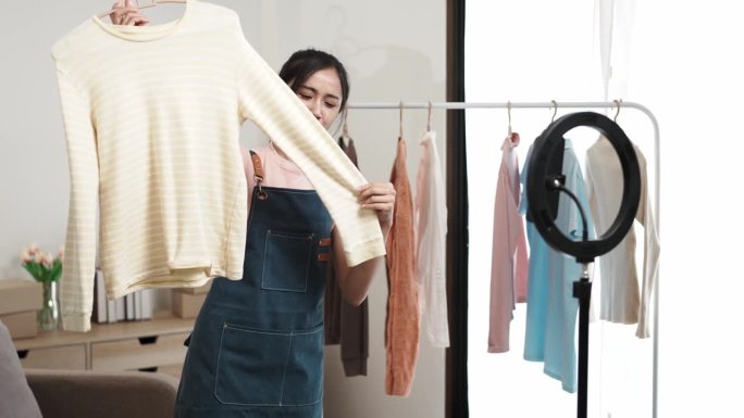 社交媒体市场概念。年轻的亚洲女卖家在移动应用程序的直播中展示二手衣服。