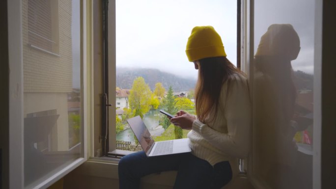 年轻女子坐在家里白色的窗台上玩智能手机。
