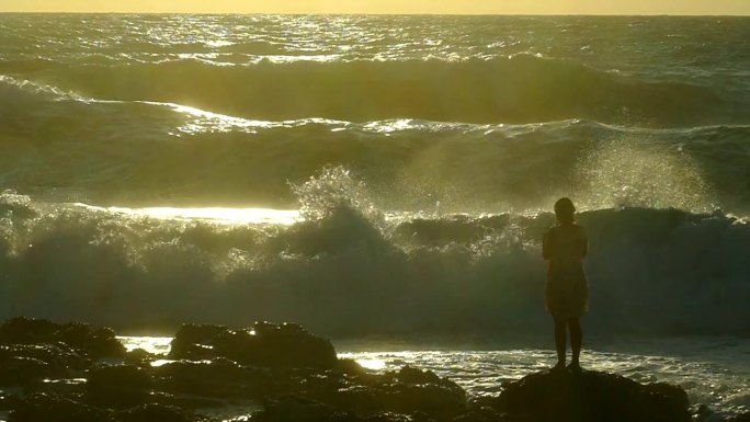 日落时，一个女孩的剪影映衬着惊涛骇浪的浪花(慢动作)
