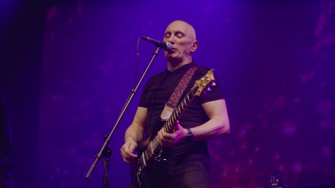 在流行乐队的演出中，贝斯吉他手在舞台上对着麦克风唱歌。舞台由蓝、粉红和红色的灯光照亮