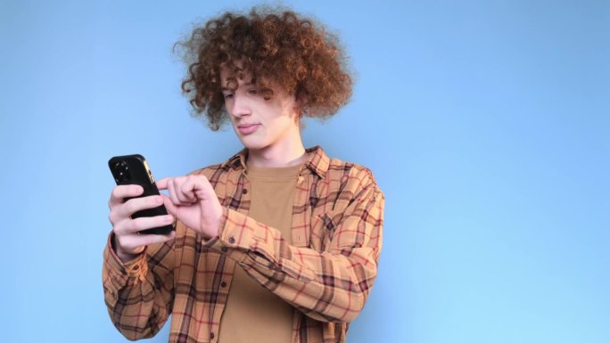 卷发红发学生站在蓝色背景上看智能手机。