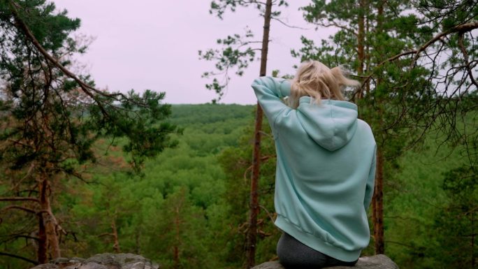 身穿淡蓝色薄荷运动衫的金发女子的后视图，对面是森林。