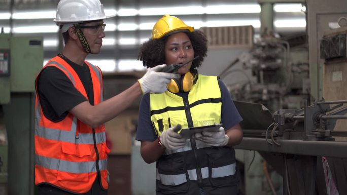 非洲裔美国工业工程师经理与亚洲修理工人机械师讨论在工业制造大型工厂中使用平板检测机器。