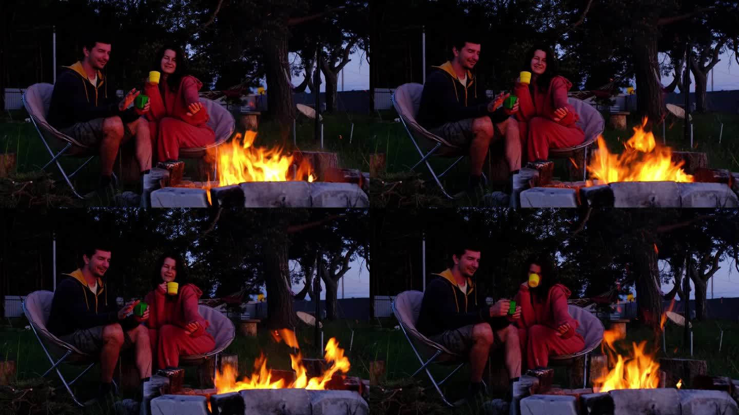 一对相爱的情侣坐在屋外院子里的营火旁的露营椅上，度过一个浪漫的夜晚。一男一女在火边暖手，喝茶