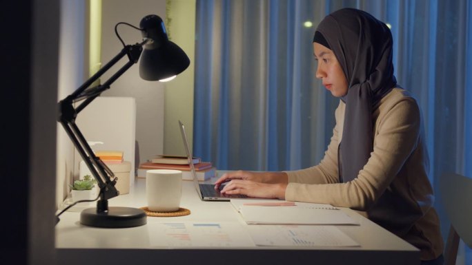 年轻快乐的亚洲穆斯林自由职业者妇女戴头巾休闲装打字键盘笔记本电脑在家里办公室加班，晚上工作。小企业主