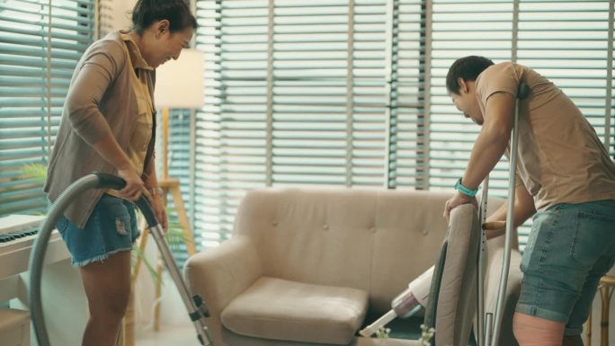 亚洲残疾夫妇在客厅忙着做家务，用吸尘器打扫房间。团队合作。例行公事。医疗保健。