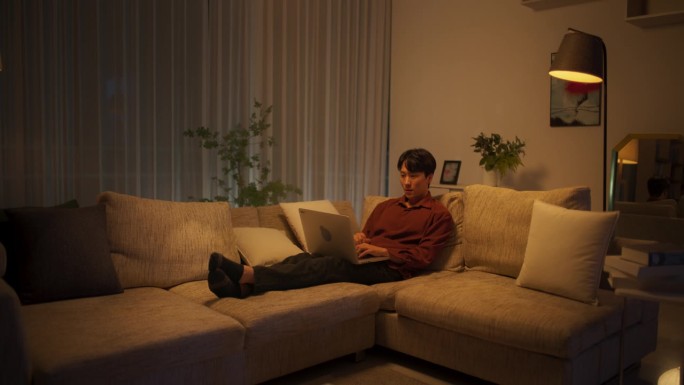 年轻英俊的亚洲男性晚上在公寓里上网，在家用笔记本电脑，在客厅的沙发上休息时在社交媒体市场上购物
