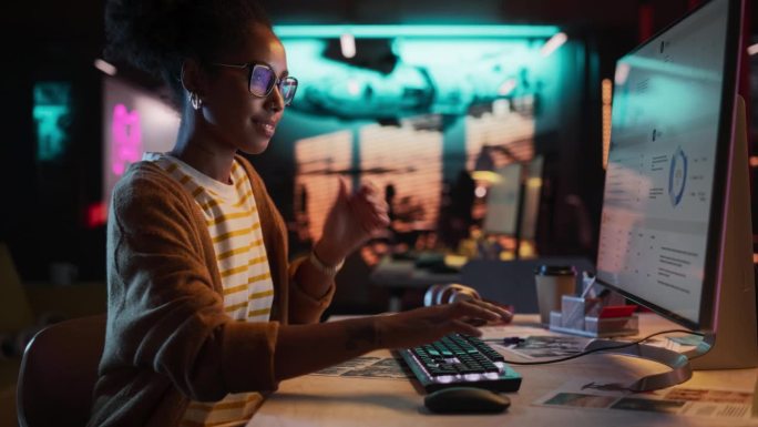 女性天才数字营销专家晚上在办公室用电脑工作。年轻的黑人女商人在创意机构，微笑和研究商业分析数据
