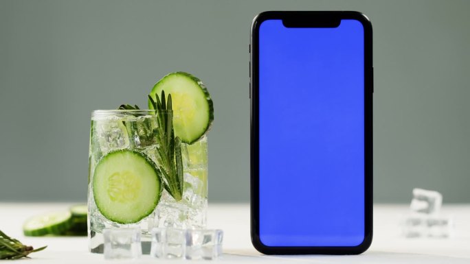 柠檬水加黄瓜和迷迭香，智能手机有蓝色色度键屏。金汤力鸡尾酒特写，灰色背景上的健康饮料。排毒饮酒，夏日