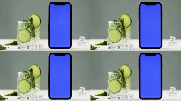 柠檬水加黄瓜和迷迭香，智能手机有蓝色色度键屏。金汤力鸡尾酒特写，灰色背景上的健康饮料。排毒饮酒，夏日