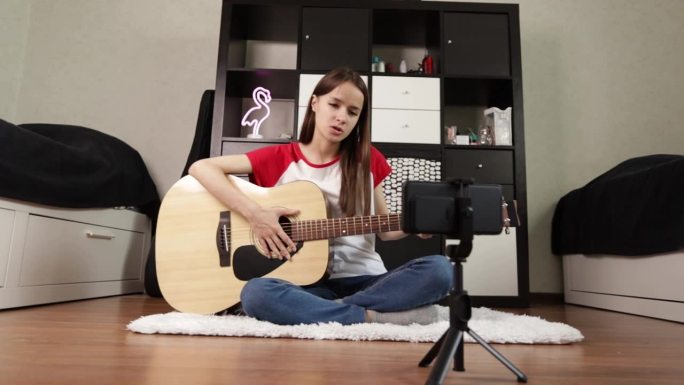 一个青少年拿着原声吉他，在智能手机上直播音乐课;一个年轻的吉他手通过博客和智能手机视频分享她的音乐之