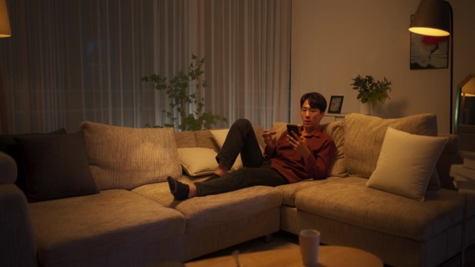 一名年轻的韩国男子在舒适的公寓里使用网上购物应用程序，用智能手机和信用卡安全地购物，更新流媒体服务的