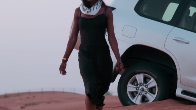 一位身穿黑色长裙、围着阿拉伯头巾、皮肤黝黑的年轻女子赤脚走在沙漠的沙地上，向一辆停在沙丘上的白色SU