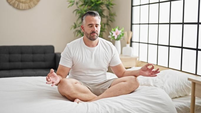 头发花白的男人坐在卧室的床上做瑜伽