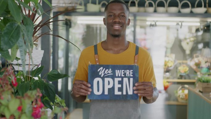 年轻的非裔美国人企业家的肖像，在新的花店里举着“是的，我们开放”的牌子