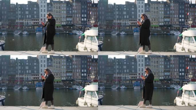 日落时分，一名女子穿着大衣，戴着太阳镜，拿着智能手机走在法国西北部卡尔瓦多斯省历史名城翁弗勒尔的海港
