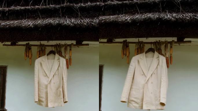 一件漂亮的白色夹克挂在衣架上，背景是一座茅草屋顶的泥木房子，屋顶下缝着一件kokuruza。垂直视频